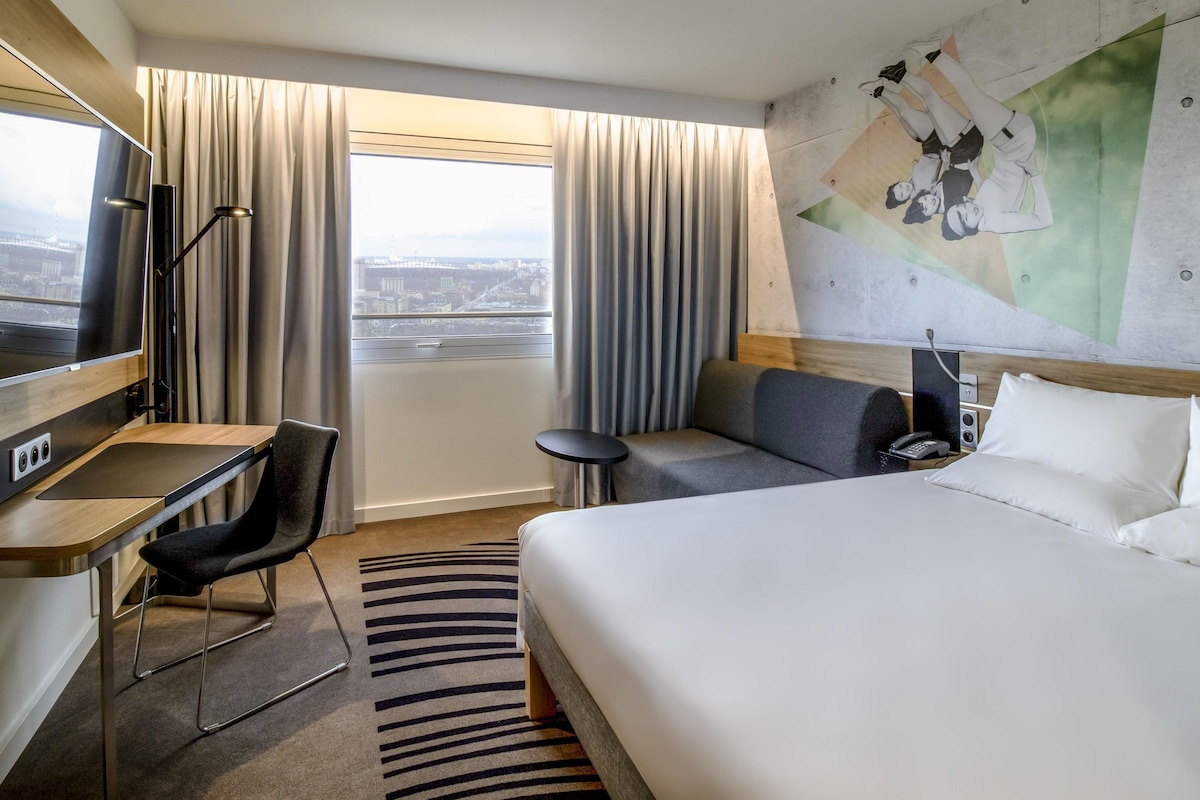 Ein Hotelzimmer mit Bett, Schreibtisch, Stuhl, Fernseher, Telefon und Blick auf die Stadt durch das Fenster.