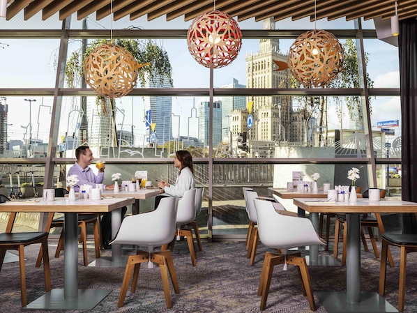 Uma sala de jantar moderna com mesas e cadeiras, grandes janelas com vista para a cidade e luminárias decorativas pendentes.