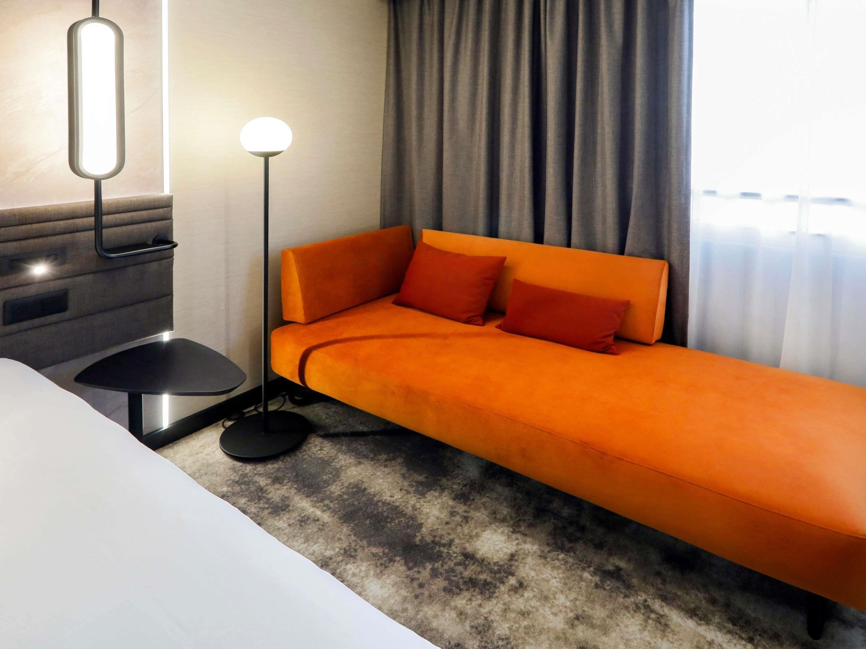 A modern hotel room with a single bed, a bedside table, a floor lamp, and a view of the outside through a window with curtains.
