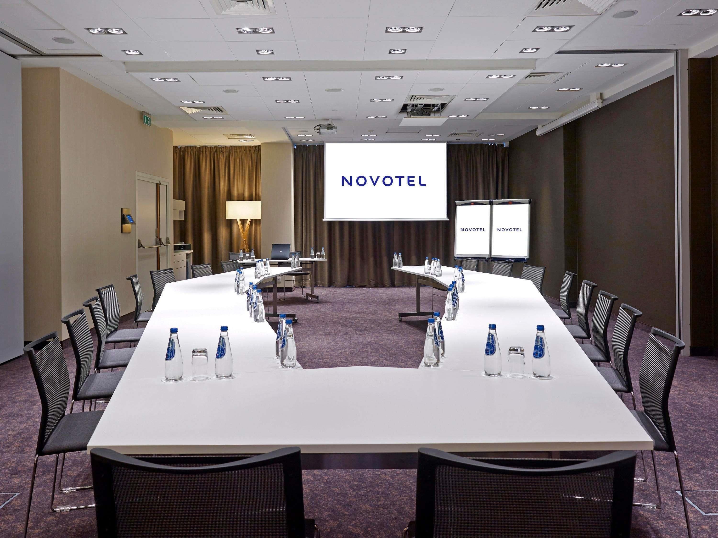 A conference room with a long table, chairs, water bottles, a laptop, and a projector screen displaying 'NOVOTEL'.