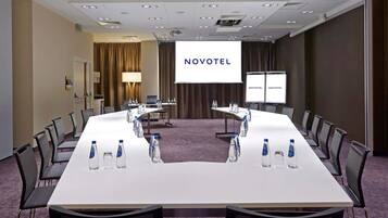 A conference room with a long table, chairs, water bottles, a laptop, and a projector screen displaying 'NOVOTEL'.