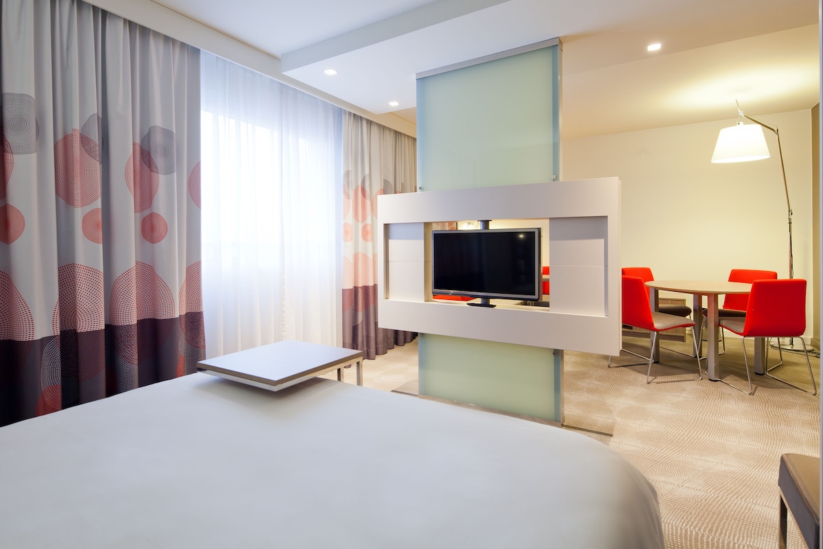 A hotel room with a bed, a TV mounted on a wall, a desk, and a chair.