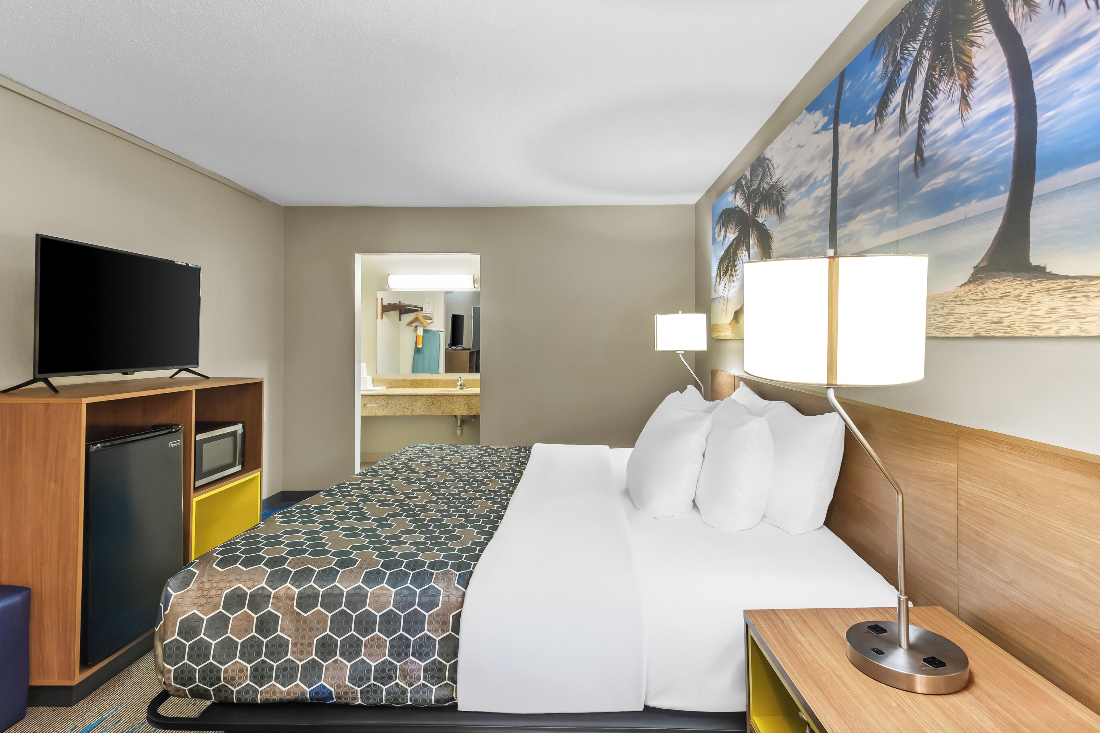 A hotel room with a bed, a desk with a television, a refrigerator, a lamp, and a painting of a beach scene.