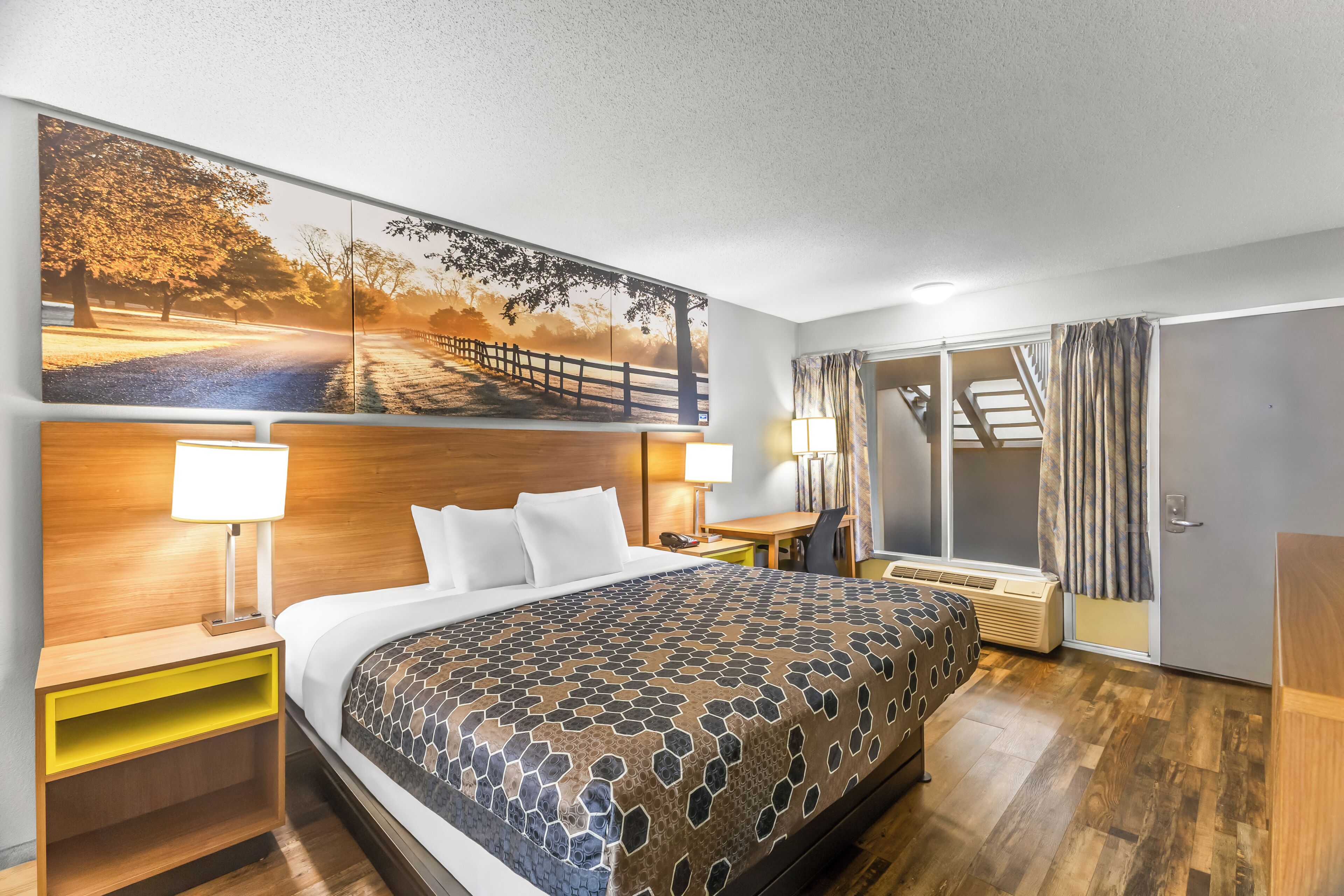 A hotel room with a large bed, a desk, a chair, a nightstand, a window with curtains, and a wall-mounted artwork.