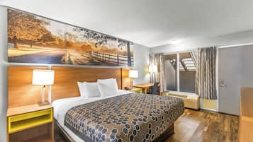 A hotel room with a large bed, a desk, a chair, a nightstand, a window with curtains, and a wall-mounted artwork.
