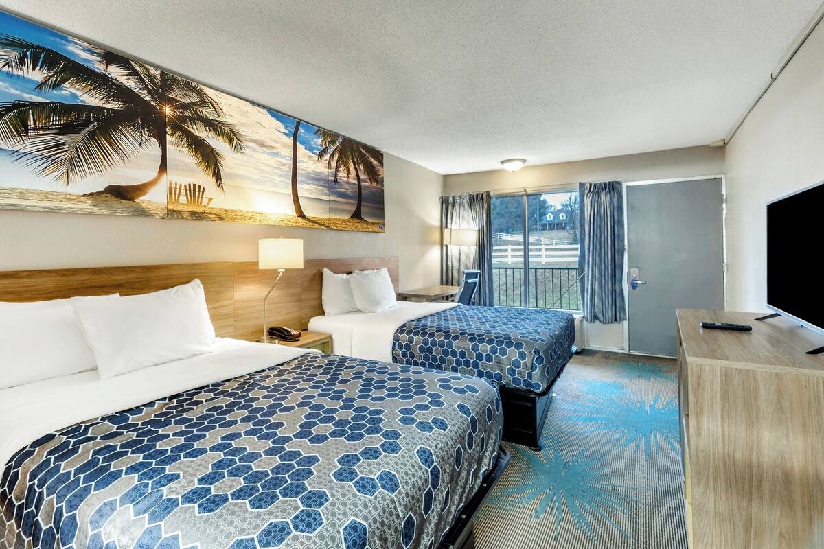 A hotel room with two beds, a TV, a desk, and a large window with curtains.