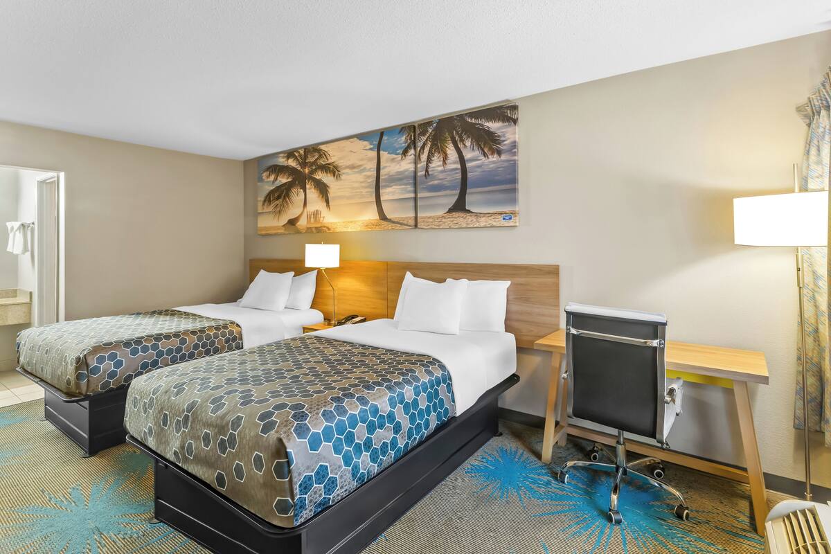A hotel room with two beds, a desk, a chair, a lamp, and a painting of palm trees.