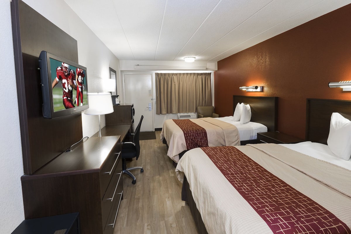 A hotel room with two beds, a desk with a chair, a TV mounted on the wall, and a window with curtains.