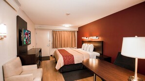 Una habitación de hotel con cama, escritorio, silla, televisión y lámpara fijada a la pared.