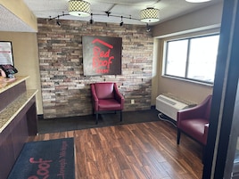 Zona de recepción con pared de piedra, una silla roja, un mostrador de recepción con quiosco digital y una ventana con vistas.