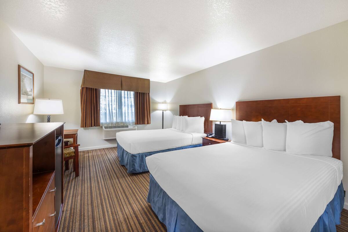 A hotel room with two beds, a desk, a chair, a lamp, and a window with curtains.