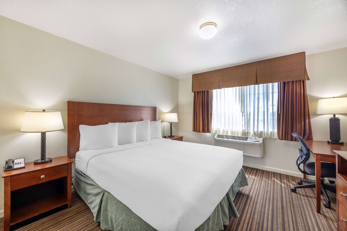 A hotel room with a large bed, a desk with a chair, a nightstand with a lamp, and a window with curtains.