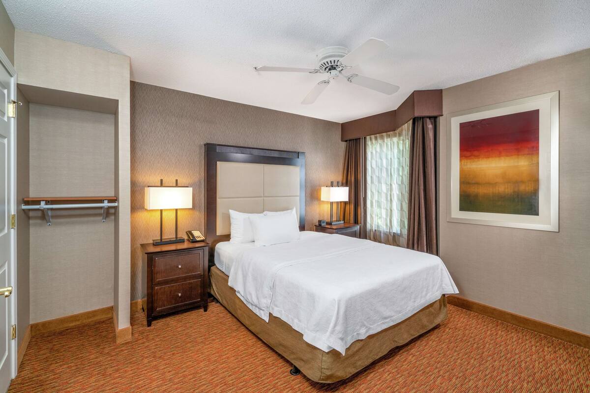 Une chambre d’hôtel avec un grand lit, deux tables de chevet avec des lampes, un ventilateur de plafond, une porte et un tableau encadré au mur.