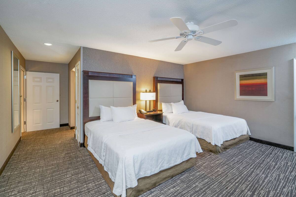 Une chambre d’hôtel avec deux lits, un ventilateur de plafond, une table de chevet avec une lampe et un tableau encadré au mur.