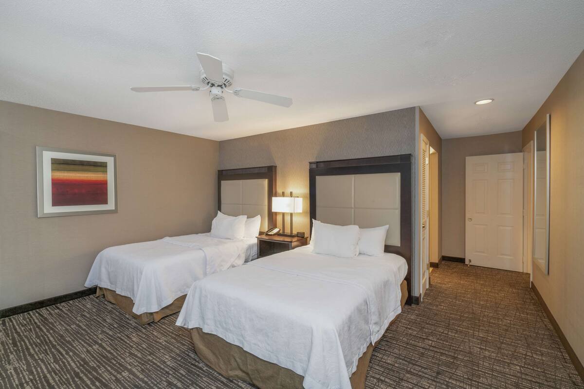 Une chambre d’hôtel avec deux lits, un ventilateur de plafond, une table de chevet avec une lampe et un tableau encadré au mur.