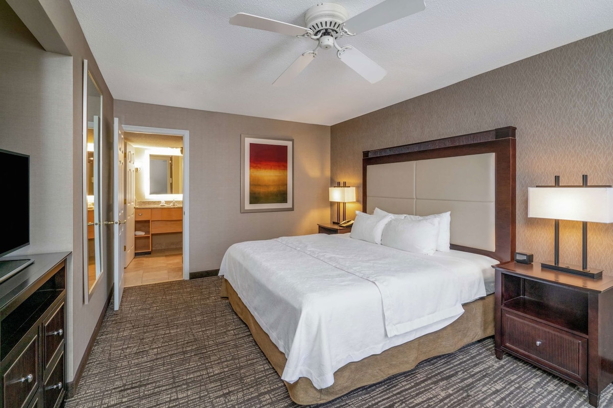 A hotel room with a bed, a television, a ceiling fan, a nightstand with a lamp, and a bathroom visible through an open door.