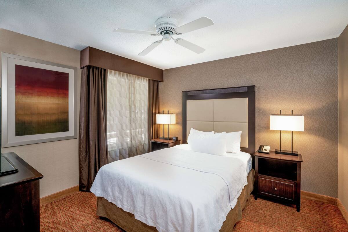 Une chambre d’hôtel avec un grand lit, deux tables de chevet avec des lampes, un ventilateur de plafond et un tableau encadré au mur.