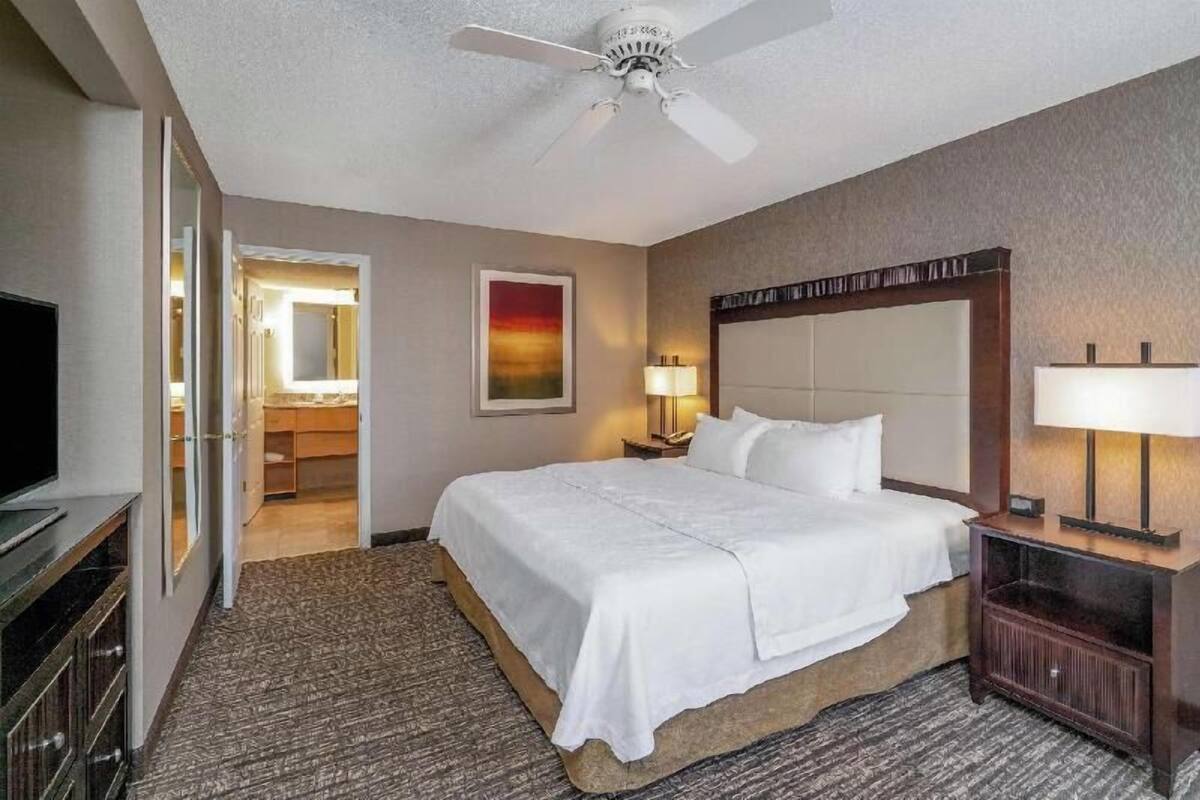Une chambre d’hôtel avec un lit, un ventilateur de plafond, une télévision, un bureau avec une lampe et un tableau au mur.