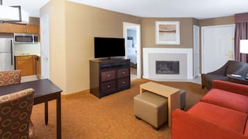 A hotel room with a kitchenette, a flat-screen TV, a fireplace, a sofa, and a dining area with a table and chairs.