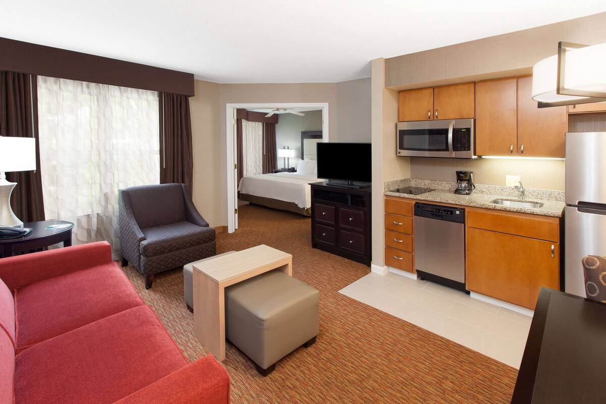 Une chambre d’hôtel avec un canapé rouge, une petite table, un téléviseur, une kitchenette équipée d’un four à micro-ondes et un lit à l’arrière-plan.