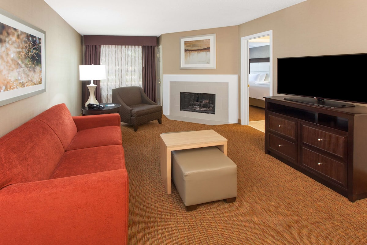 Une chambre d’hôtel avec un canapé rouge, un petit pouf, une télévision, une cheminée et un lit doté d’une tête de lit blanche.