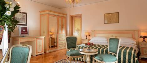 A hotel room with a bed, two chairs, a small table, a chandelier, a cabinet, and a floral arrangement.
