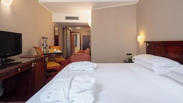 Una habitación de hotel con cama, escritorio con televisor, una silla y mesita de noche con lámpara.