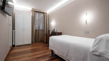 A hotel room with a wooden floor, a bed with white bedding, a wardrobe, a desk with a lamp, and a television on the wall.