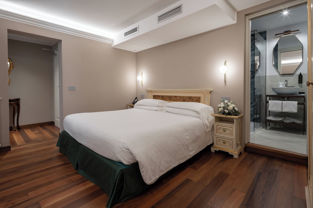 Une chambre à coucher bien aménagée, avec un grand lit, des tables de chevet, une petite table avec un vase de fleurs, et une salle de bain dotée d’une douche en verre et d’un lavabo.