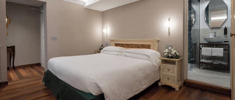 Un dormitorio ordenado con una cama grande, mesitas de noche y una mesita auxiliar con un jarrón de flores, además de un baño con ducha de vidrio y lavabo.