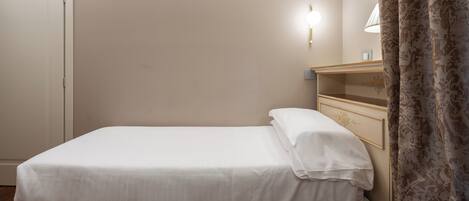 A neatly made bed with white linens in a room with a wooden floor, a wall-mounted light, and a door.