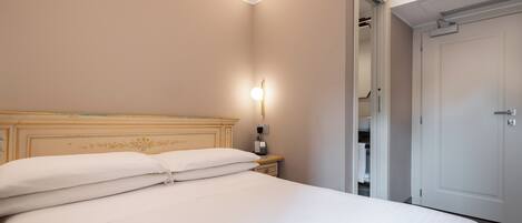 A neatly made bed with white linens, a bedside table with a lamp, and a closet with hanging clothes.