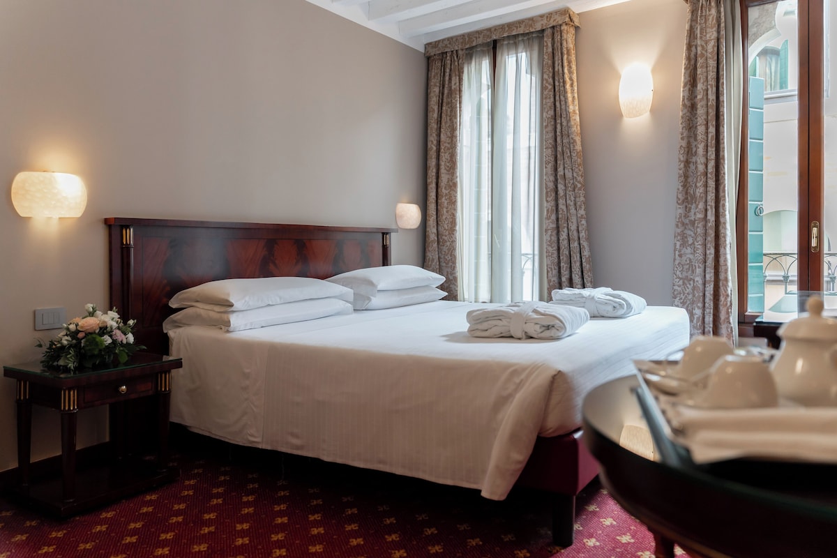 Une chambre d’hôtel avec un lit soigneusement fait, une table de chevet avec une lampe et une vue sur l’extérieur grâce à de grandes fenêtres.