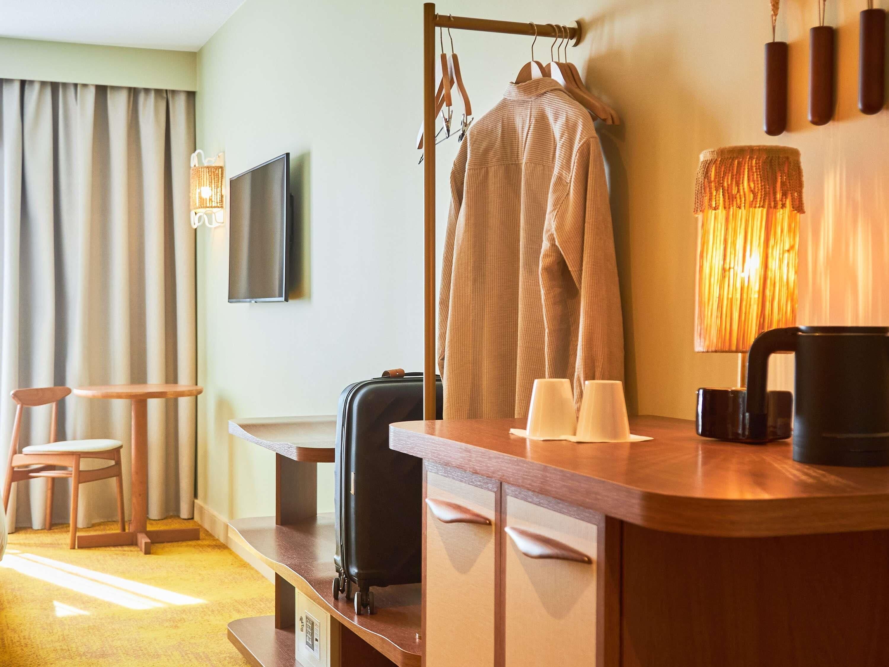 A hotel room with a wooden desk, a television, a coat rack with a coat, a suitcase, and a lamp.