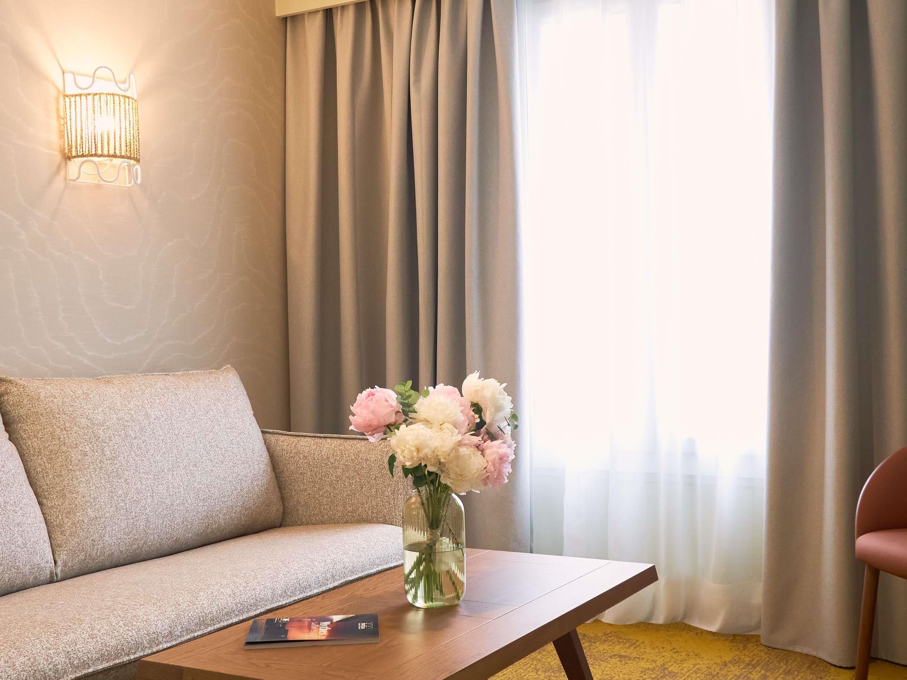 A hotel room with a sofa, a wooden table with a vase of flowers, a lamp, and a view of the outdoors through curtains.