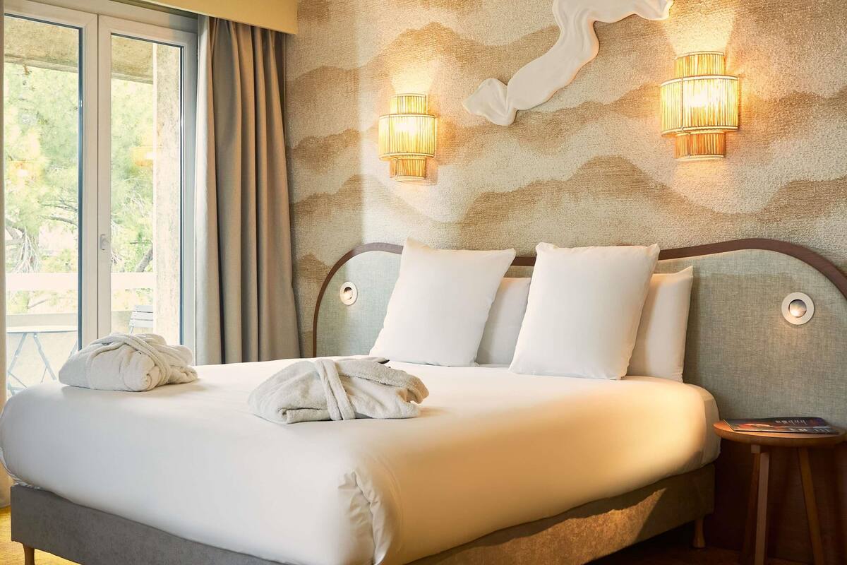 Une chambre d’hôtel avec un lit, deux lampes fixées au mur, une fenêtre avec des rideaux et une vue sur la verdure à l’extérieur.