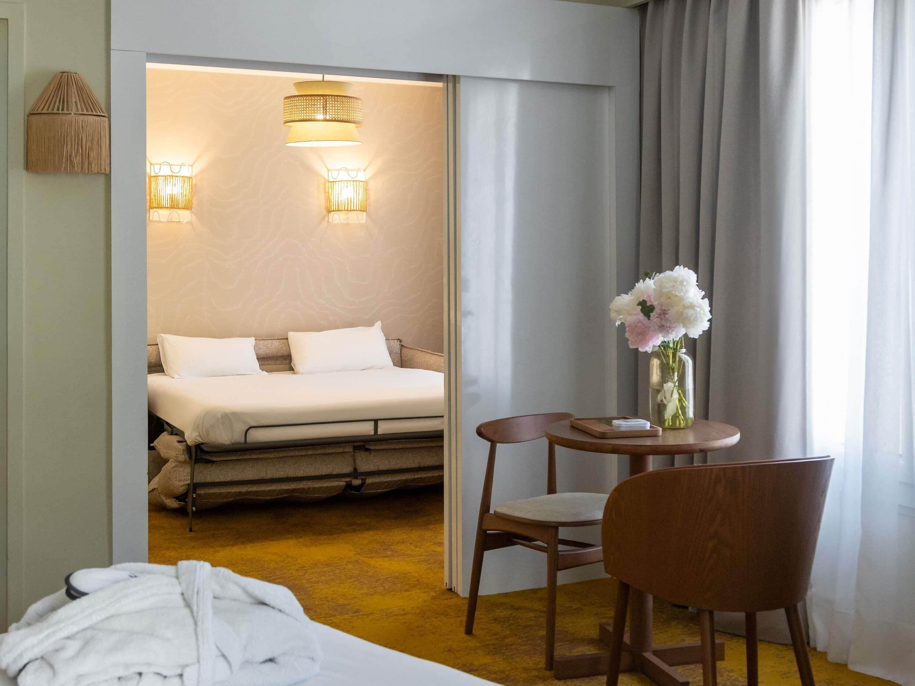 A hotel room with a bed, a chair, a table with a vase of flowers, and two wall-mounted lamps.