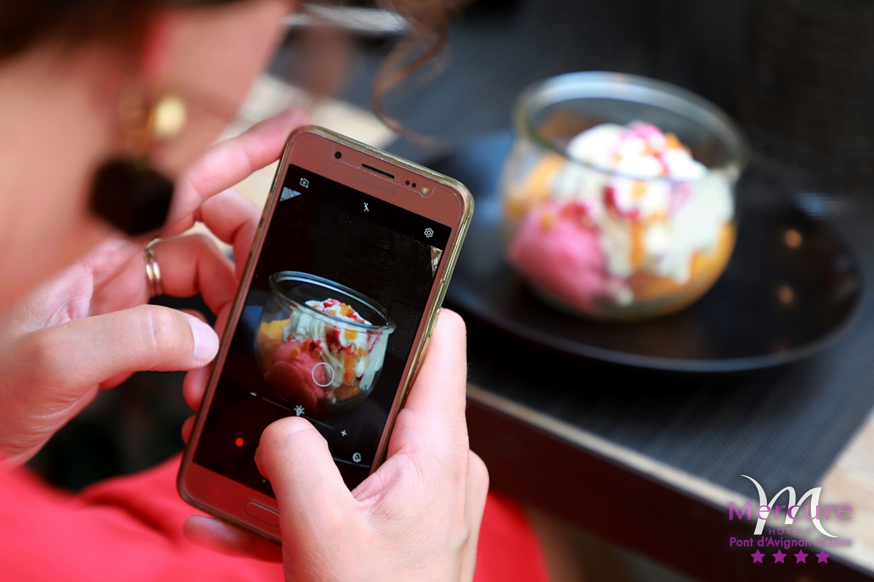 A person holding a smartphone with a dessert in a glass container on a table.