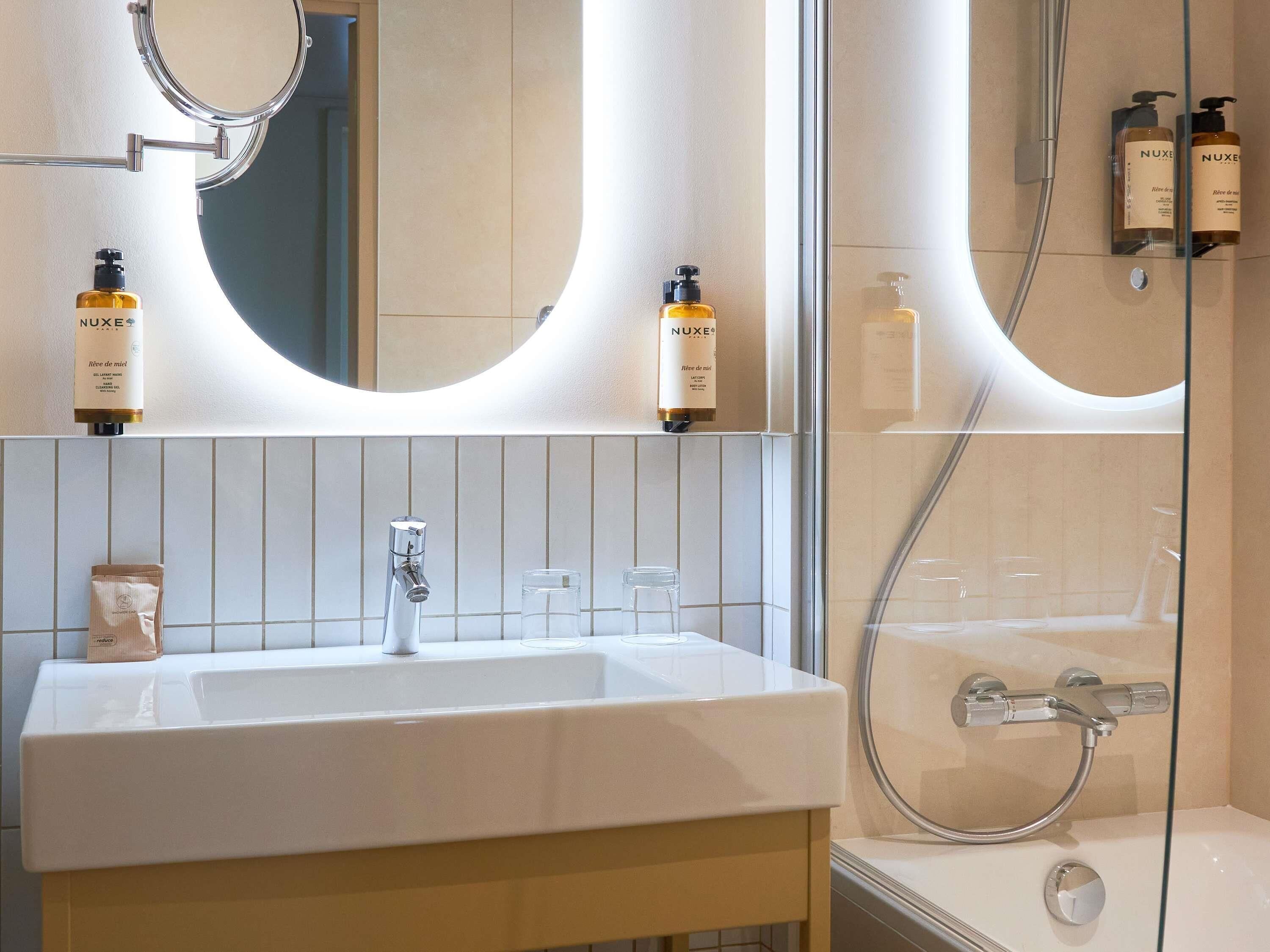 A bathroom with a white sink, a mirror, and two Nuxe shower bottles.