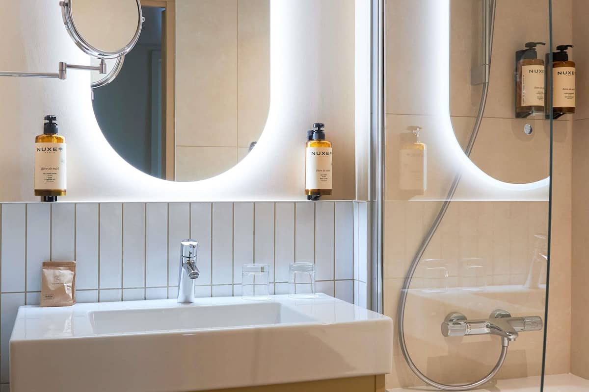 A bathroom with a white sink, a mirror, and two Nuxe shower bottles.