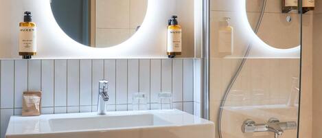 A bathroom with a white sink, a mirror, and two Nuxe shower bottles.