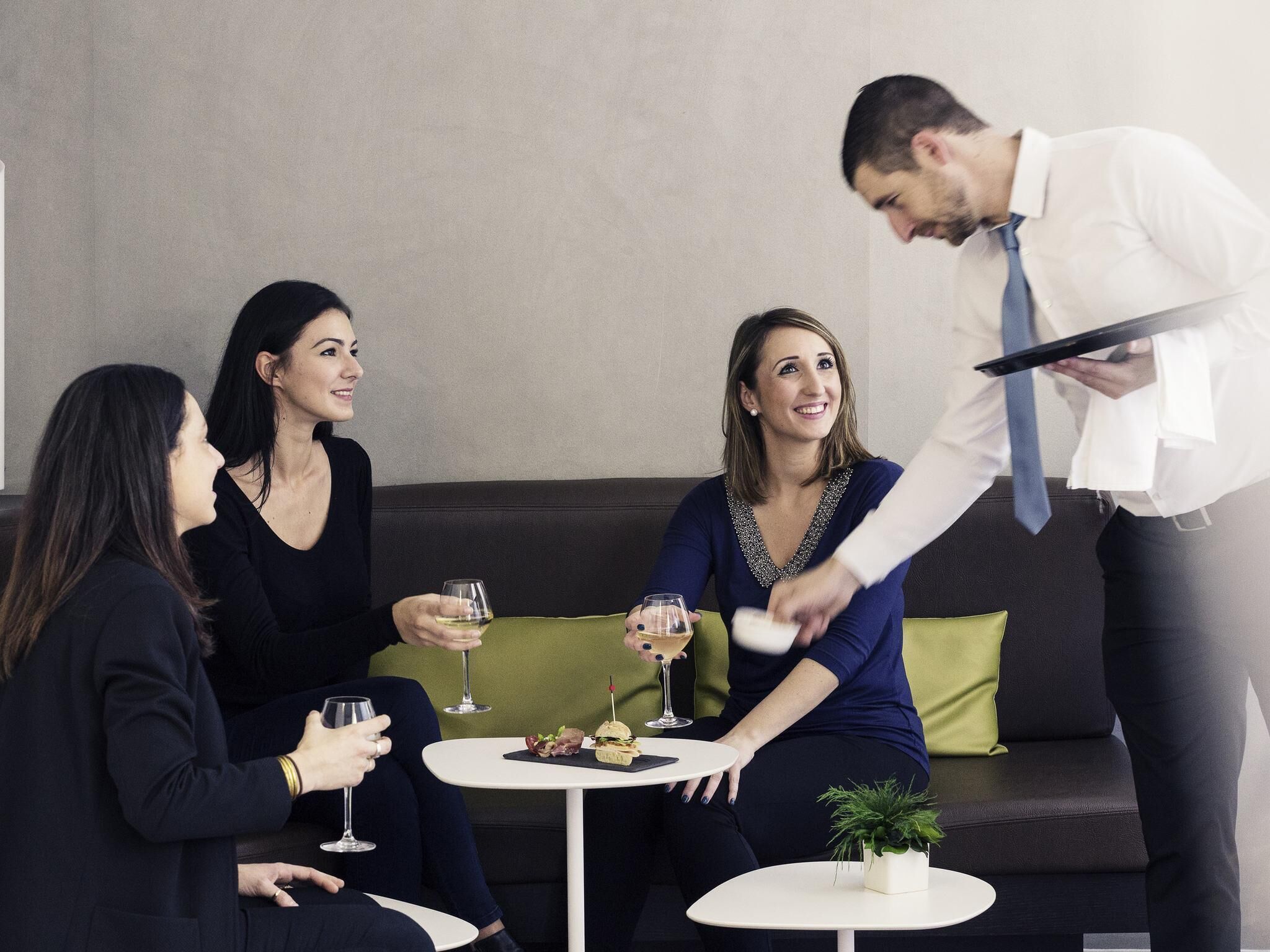 A group of people seated around a table with drinks, a waiter serving, and a small plant on the table.