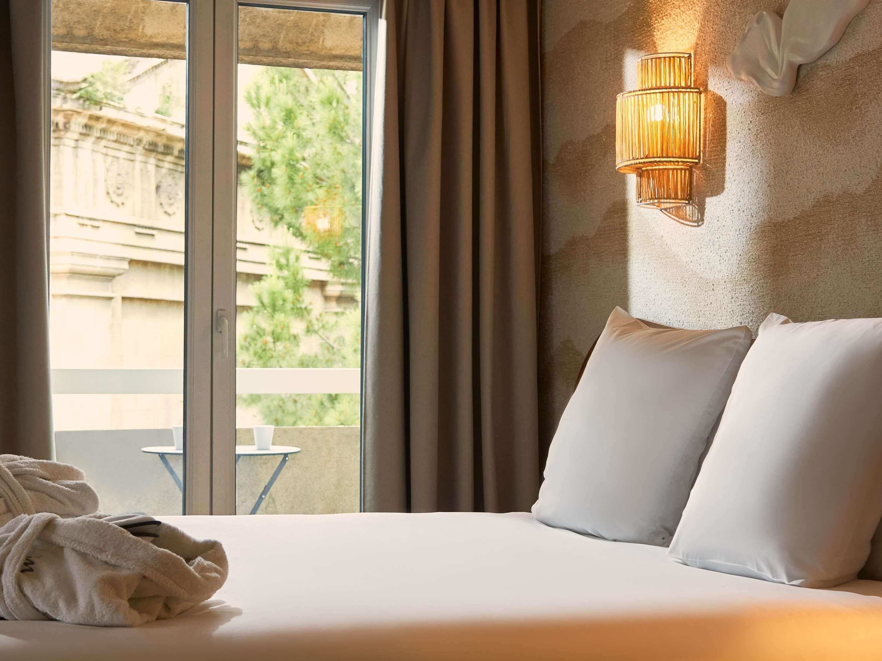 A hotel room with a large window, a bed with white linens, a wall-mounted lamp, and a view of greenery outside.
