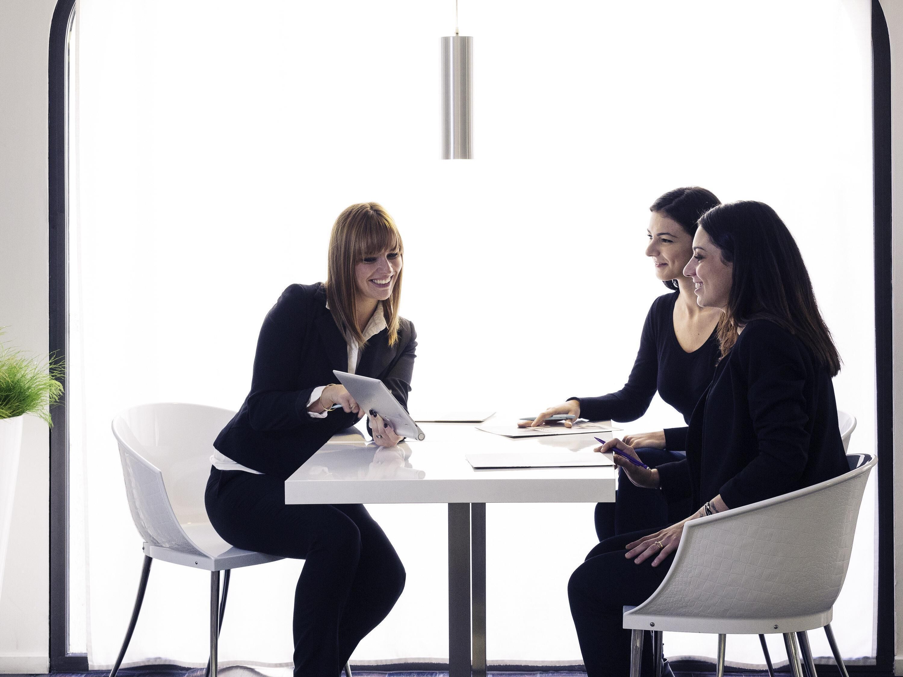 Three individuals in a meeting room with a white table, chairs, and a hanging light fixture.