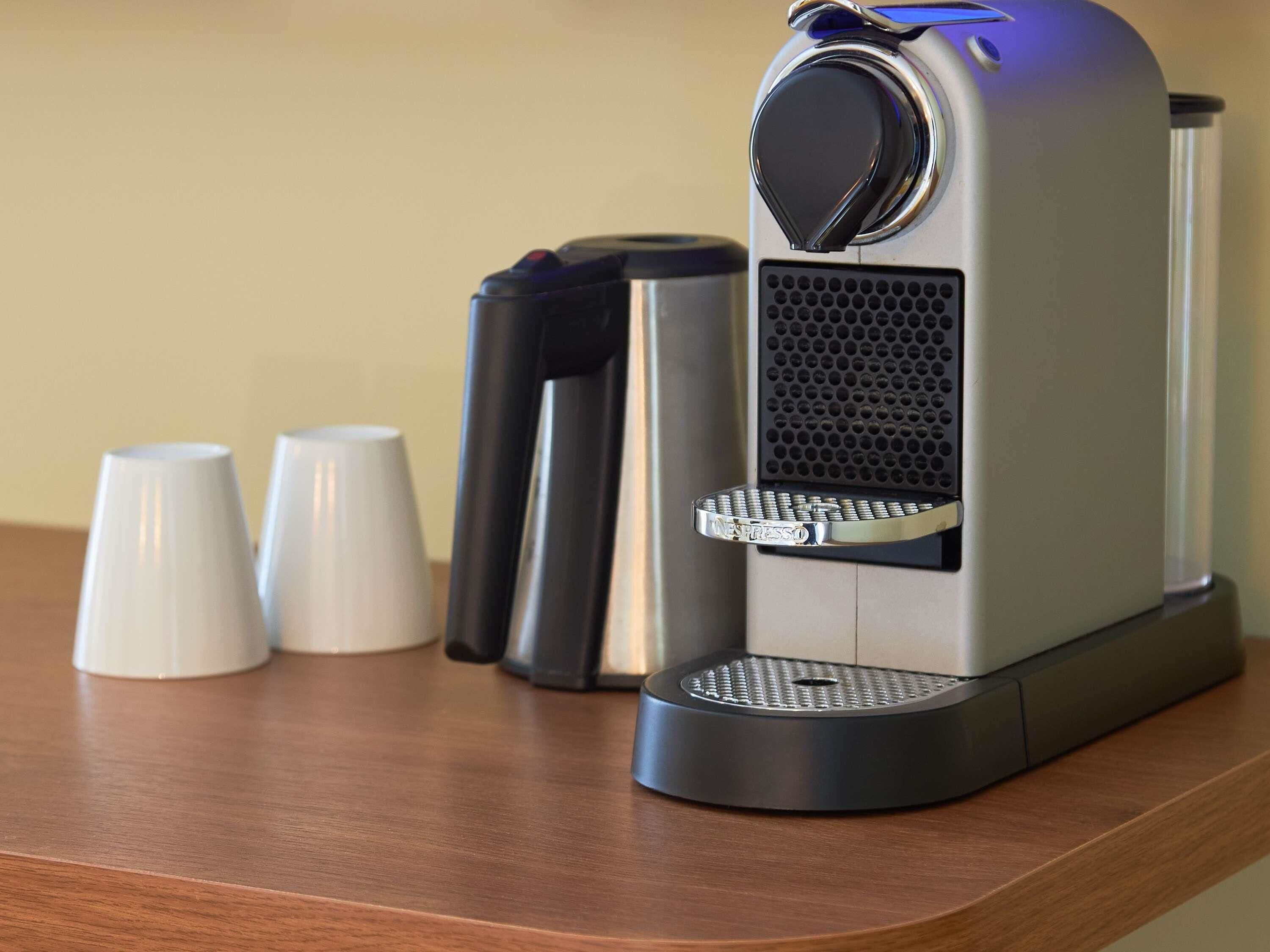A coffee machine with a drip tray, two white cups, and a black container on a wooden surface.