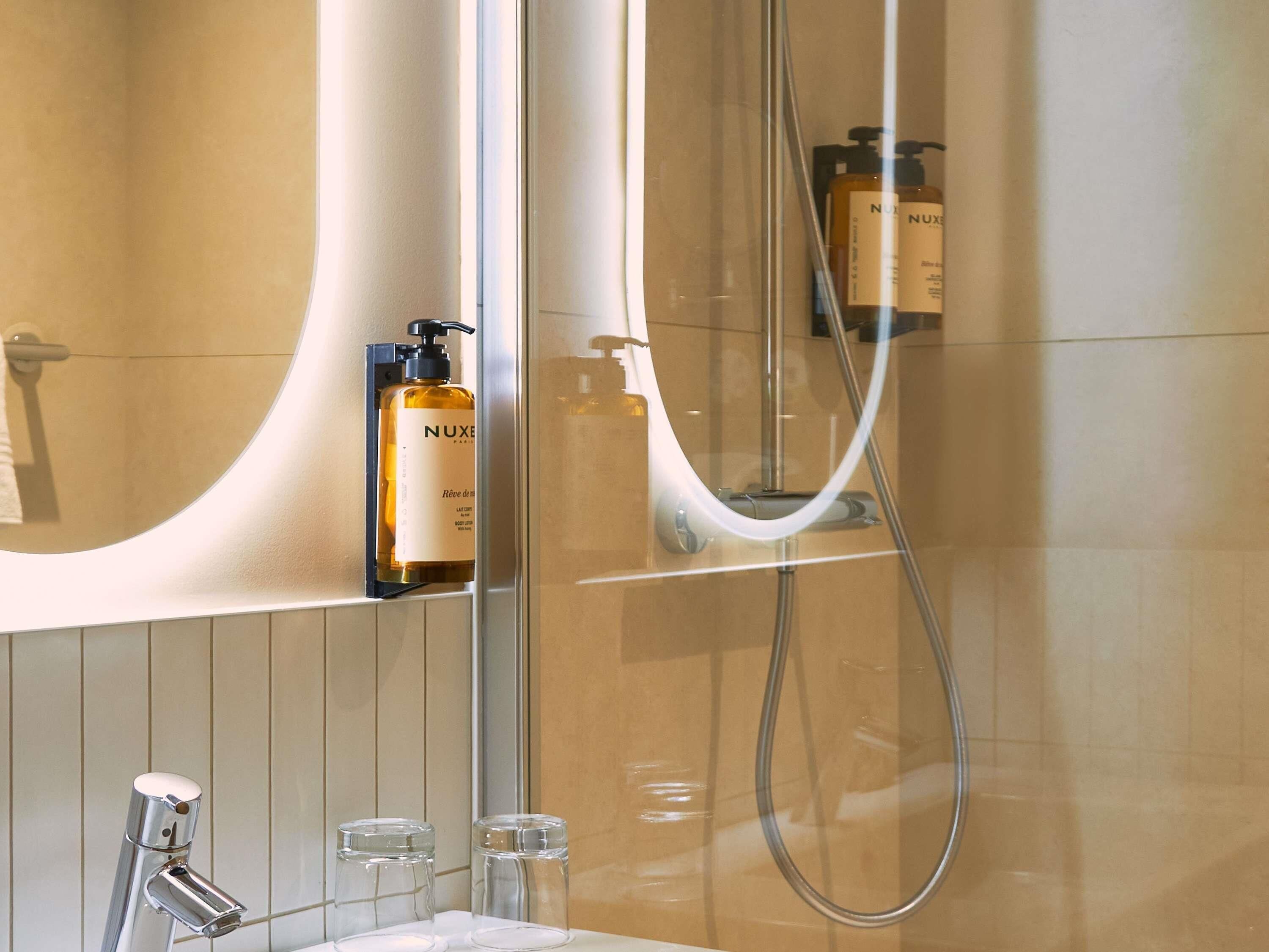 Ein Badezimmer mit Spiegel, Waschbecken, Dusche und einer Flasche Nuxe Körperlotion.