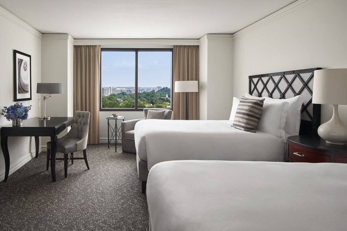 Habitación de hotel con dos camas, un escritorio, una silla, una lámpara y vista de la ciudad a través de un ventanal grande.