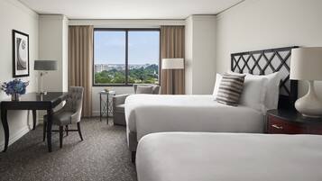 A hotel room with two beds, a desk, a chair, a lamp, and a view of the city through a large window.