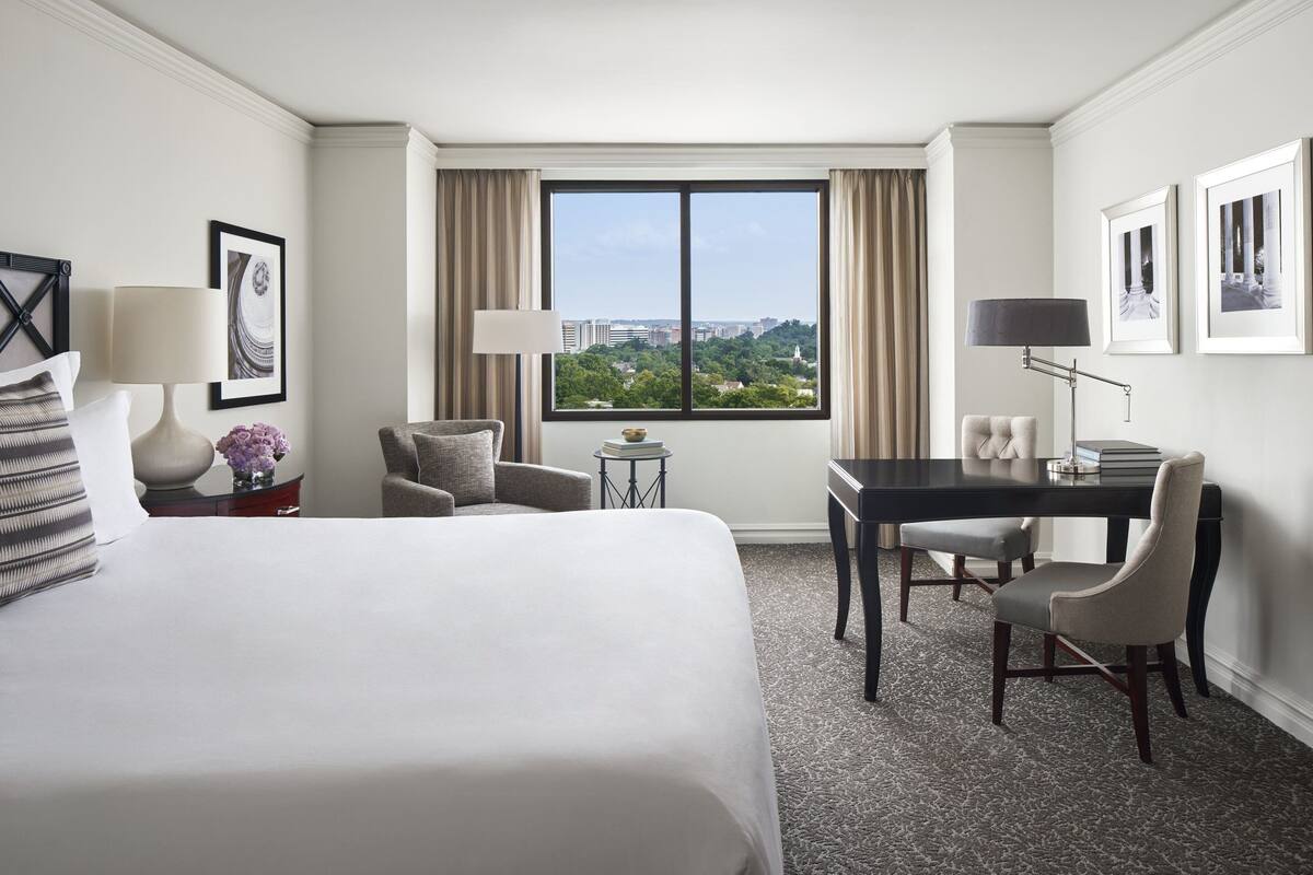 Habitación de hotel con una cama grande, un escritorio, dos sillas, una mesita y una ventana con vista a la ciudad.