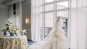 A wedding dress in a room with large windows, a table with floral arrangements, and a patterned carpet.